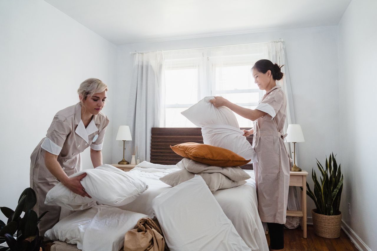 Duas profissionais da governança preparando a cama de um quarto de hotel